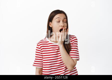 Jeune femme fatiguée bâillant, se sentant épuisée ou endormie, se réveillant tôt le matin, debout dans un t-shirt rouge sur fond blanc Banque D'Images