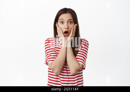 Portrait d'une femme brunette choquée, qui se flatte, prend la mâchoire et regarde, tenant les mains sur le visage surpris, regardant le sourire inquiétant, se tenant sur le blanc Banque D'Images