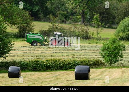 Production de foin ou d'ensilage (agriculteur travaillant dans un tracteur agricole sur une ramasseuse-presse à balles pour le travail dans les champs ruraux, collecte de l'herbe sèche et balles rondes enveloppées - Yorkshire England, Royaume-Uni. Banque D'Images