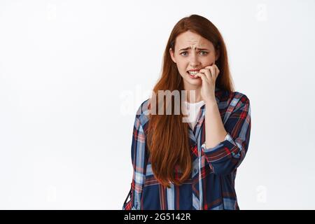 Une fille à tête rouge effrayée semble anxieux, mordant les ongles et regardant effrayée, paniquant, se sentant inquiète ou nerveuse, se tenant sur fond blanc Banque D'Images