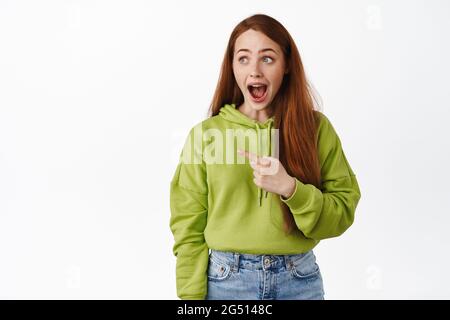 Surprise et heureuse fille de gingembre lâcher mâchoire excitée, pointant et regardant gauche dans la crainte, en regardant la publicité de shopping, se tenant sur fond blanc Banque D'Images