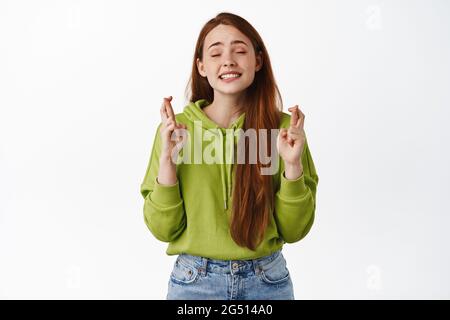 Image d'une fille à tête rouge pleine d'espoir, croiser les doigts et souhaiter, piquer les yeux et les yeux, anticiper les bonnes nouvelles positives, se sentir chanceux, blanc Banque D'Images