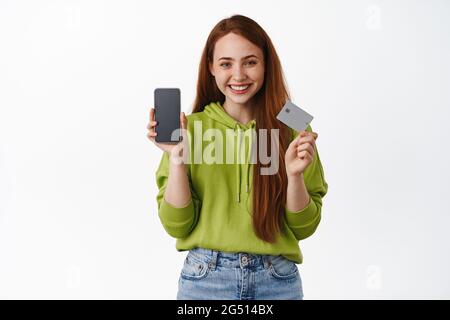 Achats en ligne. Une fille souriante montre une interface d'application mobile et une carte de crédit, des boutiques dans un magasin Internet, achetant sans contact à la maison, se tenant contre Banque D'Images