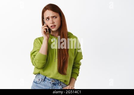 Agacé redhead fille parle au téléphone, ayant la conversation désagréable de confusion, appelant quelqu'un sur le téléphone cellulaire, fond blanc Banque D'Images