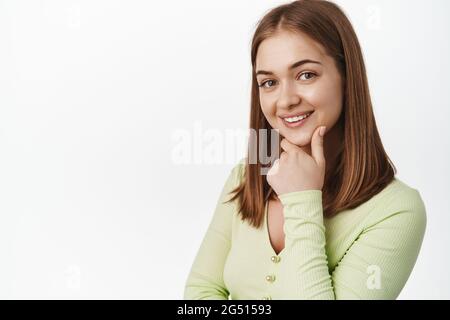 Gros plan de jeune fille confiante look intrigué, intéressé par le sourire, le menton de toucher et le sourire réfléchi, regardant la caméra réfléchir, debout Banque D'Images