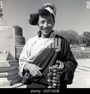 Années 1950, photo historique par J Allan Cash d'un jeune souriant, heureux debout à l'extérieur du temple bouddhiste, mulagandhakuti vihara, Sarnath, Varanasi, inde, tenant des guirlandes de fleurs ou des tresses, qui sont portés autour du cou et qu'il offre ou vend aux visiteurs. Le temple a été construit en 1931 - sur le site où le Bouddha Gautama a médié pendant sa première saison des pluies - par Angarika Dharmapala et est maintenu et dirigé par la société Mahabodhi dont il a fondé. Banque D'Images