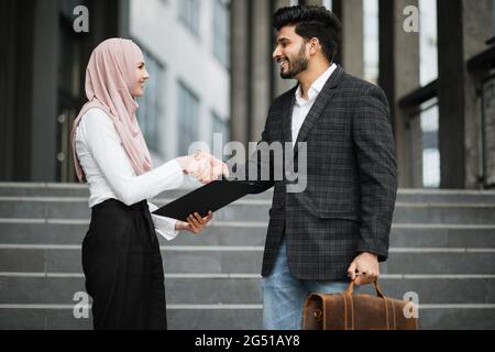 Beau homme hindou et charmante femme dans hijab en serrant les mains et en souriant en se tenant sur les escaliers près du bâtiment de bureau. Coopération réussie de deux partenaires. Banque D'Images