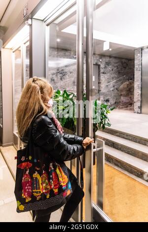 Femme blonde portant un masque facial et un sac à main froid ouvrant la porte de son appartement. Banque D'Images