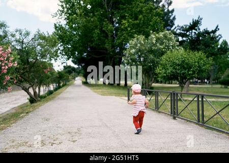 Petite fille au panama marche le long de la route dans un parc vert parmi les arbres en fleurs Banque D'Images