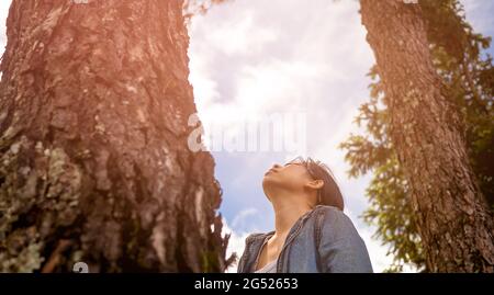 Une femme heureuse se tient près d'un arbre majestueux et regarde au sommet en profitant de la nature dans le jardin de printemps. Concept de voyage et de non-déforestation. Banque D'Images