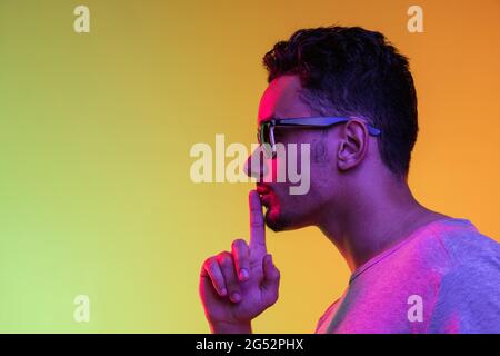 Portrait du jeune homme de Latina dans des lunettes de soleil se posant isolé sur fond vert jaune dégradé en lumière néon. Vue latérale Banque D'Images