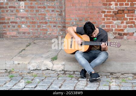 jeune garçon latin appréciant jouer de la guitare tout en étant assis sur le sol avec un fond de mur de brique, souterrain, urbain Banque D'Images