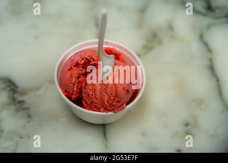 Vue en hauteur d'une cuillère à usage unique en plastique blanc dans un bol en polystyrène expansé rempli de pelles à crème glacée à la fraise sur une table en marbre. Banque D'Images