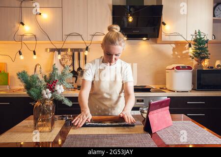 Une femme suit sa recette sur une tablette numérique et roule la pâte avec une broche pour cuire des biscuits de pain d'épice de noël dans sa cuisine. Banque D'Images