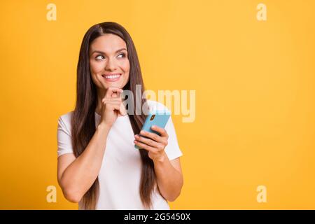 Portrait photo d'une femme curieuse et attentiste souriant en touchant le menton en maintenant le téléphone portable isolé sur fond jaune vif Banque D'Images