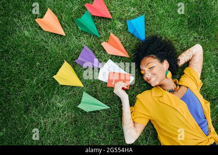 Concept de déplacement. Femme multi-ethnique avec des billets d'avion et un passeport international à la main rêve de vacances allongé sur l'herbe verte autour de Pape coloré Banque D'Images