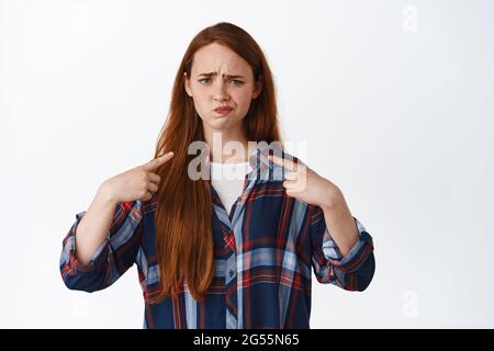 Sceptique redhead femme se pointant vers elle, grimaquant et frowning déçu, étant incertain dans elle, mécontente, debout sur fond blanc Banque D'Images