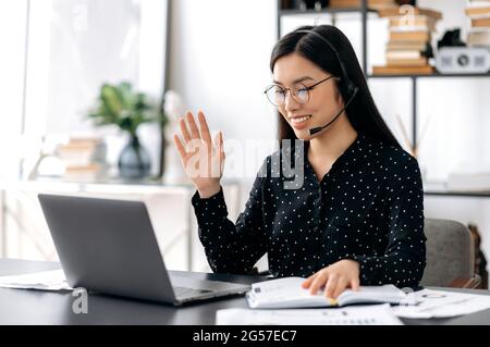 Une jeune femme asiatique agréable et intelligente portant des lunettes et un casque, une personne indépendante, un employé de soutien ou un opérateur de ligne directe s'assoient à un bureau, parle au client, mène une consultation, agite la main, sourit Banque D'Images