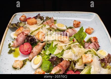 Salade César aux herbes, œufs de bacon de poulet et tomates sur une assiette carrée claire, sur fond sombre Banque D'Images