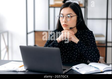 Bouleverser la jeune femme brune asiatique avec des lunettes et un casque, un opérateur ou un responsable de soutien, assis à un bureau, regardant de côté, malheureuse, éprouvant du stress, a besoin de repos, inquiet Banque D'Images