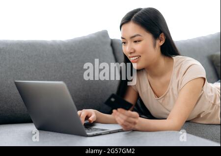 Achats en ligne. Positive belle jeune femme asiatique brunette couché sur un canapé, utilise un ordinateur portable et une carte de crédit pour payer pour les achats et la livraison en ligne, saisit des données, regarde l'écran, sourit Banque D'Images