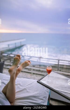 Les pieds des femmes et un cocktail dans la chambre d'hôtel avec vue sur la mer au lever du soleil Banque D'Images