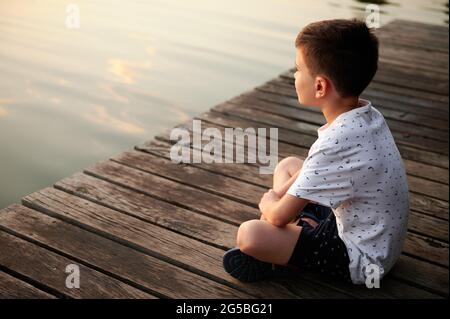 Un écolier serein assis sur un quai et profitant de la tranquillité de la nature tout en regardant le paysage du lac au coucher du soleil Banque D'Images