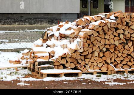 Bois de chauffage fraîchement coupé soigneusement empilé sur des palettes en bois recouvertes de neige tombant devant la maison familiale de banlieue le jour froid du printemps Banque D'Images