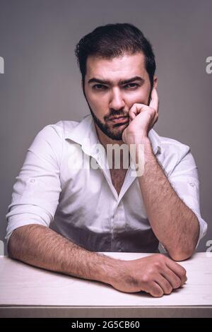 Homme d'affaires sérieux avec une barbe assise à table et un visage perplexe. Guy en chemise blanche avec expression sceptique sur fond gris en studio Banque D'Images