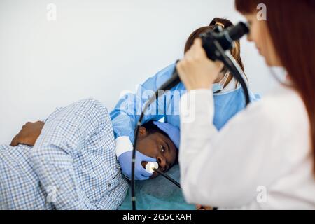 Jeune homme afro-américain patient allongé sur le canapé à l'hôpital, pendant l'endoscopie, tandis que l'équipe de deux femmes caucasiennes médecins effectuant la procédure Banque D'Images