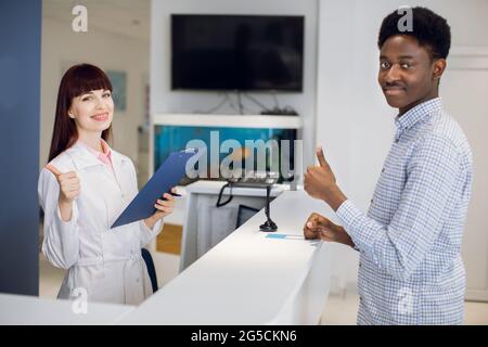 Une femme médecin consulte une patiente afro-américaine masculine dans une clinique médicale moderne. Le patient et le médecin se tenant près de la réception et montrant le pouce vers le haut Banque D'Images