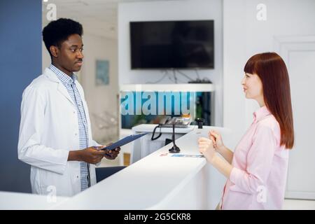 Belle jeune femme patiente parle avec un médecin afro-américain administrateur tenant le presse-papiers et souriant, debout près de la table dans la clinique Banque D'Images