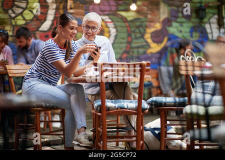 caucasain femme âgée et sa jeune amie femme regardant quelque chose ensemble sur un téléphone cellulaire, souriant. mère et fille adulte conce relation Banque D'Images
