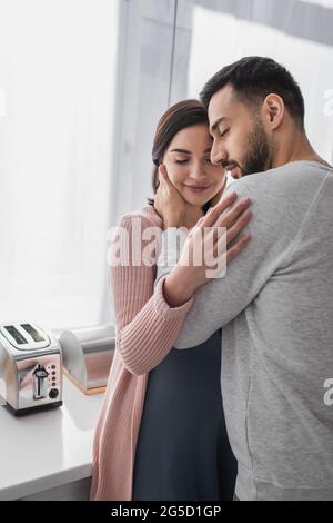 jeune homme heureux embrassant une femme enceinte avec les yeux fermés dans la cuisine Banque D'Images