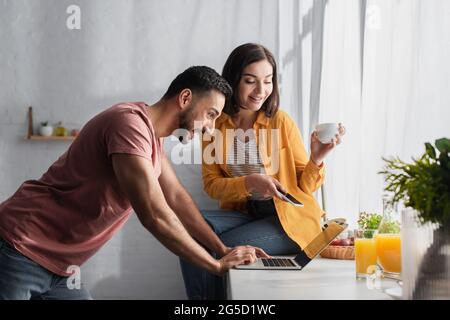 jeune femme souriante assise avec un téléphone portable et une tasse de café près d'un petit ami avec un ordinateur portable à la maison Banque D'Images