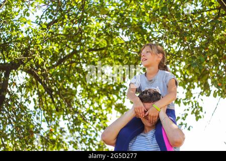 Frère défoqué de la sœur de circonscription à l'arrière. Portrait d'une fille heureuse sur les épaules de l'homme, porcgyback. Petite mouche. Famille jouant à l'extérieur. Arbre vert flou Banque D'Images