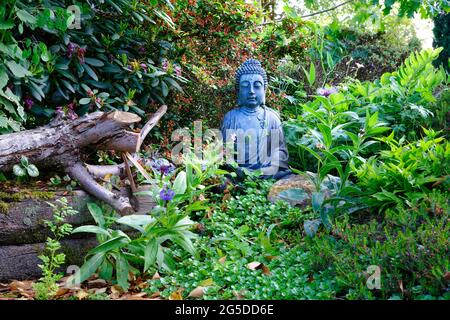 Figurine Bouddha assise médiante, située dans le jardin de feuilles vertes et de petites fleurs avec des rochers et de vieilles branches Banque D'Images