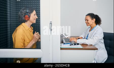 Examen audiologique. Audiologiste effectuant un contrôle auditif auprès d'une femme âgée dans un kiosque audiométrique insonorisé. Traitement de la perte auditive, vue latérale Banque D'Images