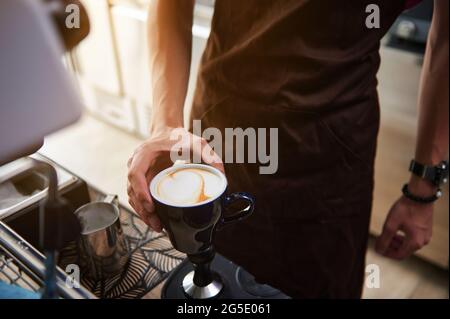 Gros plan. Tasse avec cappuccino dans la main du barista masculin. Barista derrière un comptoir de bar dans un café Banque D'Images