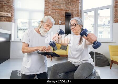 Charmante femme âgée s'entraîner avec des haltères sur le fitball et regarder le tutoriel sur la tablette qui tient son mari entre les mains. Concept de retraite, gadgets modernes et d'entraînement. Banque D'Images