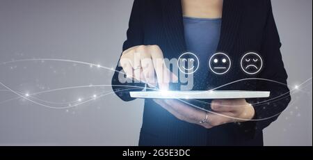 Score de visage souriant pour un sondage de satisfaction. Tablette blanche en main femme d'affaires avec hologramme numérique visage signe d'émotion sur fond gris. Client Banque D'Images
