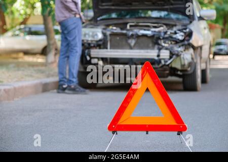 Triangle rouge d'avertissement d'accident de voiture sur la route devant la voiture de démolition. Homme inspectant la voiture de naufrage après un accident. Le conducteur regarde sous le capot de Banque D'Images