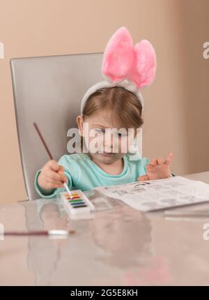 Une petite fille dans un joli serre-tête avec des oreilles de lapin roses tire avec un pinceau avec des peintures à l'aquarelle Banque D'Images