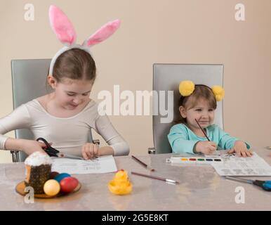 Les sœurs drôles dans les bandeaux drôles avec pompompons jaunes et oreilles de lapin rose peignent et découpez les décorations de Pâques du papier Banque D'Images