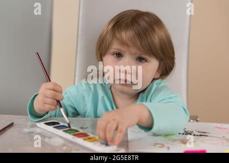 Une fille de trois ans dans un chemisier turquoise à la table dessine avec des peintures à l'aquarelle. Il regarde la caméra. Portrait Banque D'Images