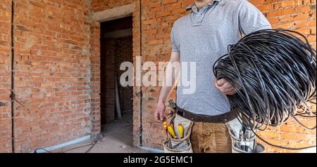 Un électricien dans un casque de sécurité regarde le mur tout en tenant un câble électrique. Banque D'Images