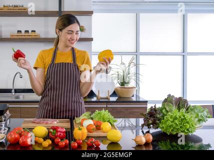 La jeune femme de ménage se tient souriante, tient le poivron rouge et jaune avec les deux mains. En regardant le jaune sur la gauche. Le comptoir de cuisine plein de va Banque D'Images