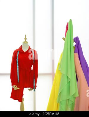 La chemise rouge et le mètre ruban sont sur le mannequin. Un porte-vêtements avec des tissus colorés devant la grande fenêtre. Atmosphère matinale dans la queue Banque D'Images