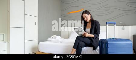 Femme d'affaires asiatique en costume noir assise avec un ordinateur tablette dans la main. Ses bagages ont été placés à côté du lit. Atmosphère de travail à l'extérieur pendant le Banque D'Images