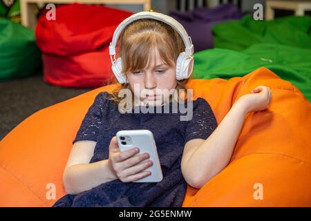 La petite fille utilise le téléphone et écoute de la musique avec un casque tout en étant assise sur un fauteuil à sac. Banque D'Images